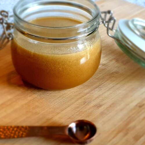 Glass jar filled with homemade caramel sauce on a wooden surface, with a metal measuring spoon in the foreground.