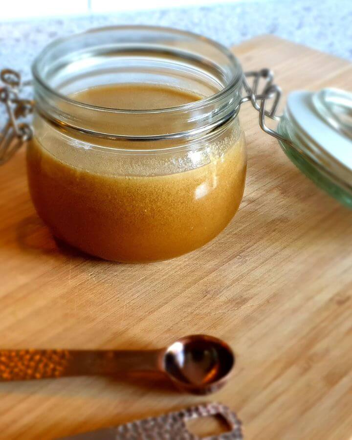 Glass jar filled with homemade caramel sauce on a wooden surface, with a metal measuring spoon in the foreground.