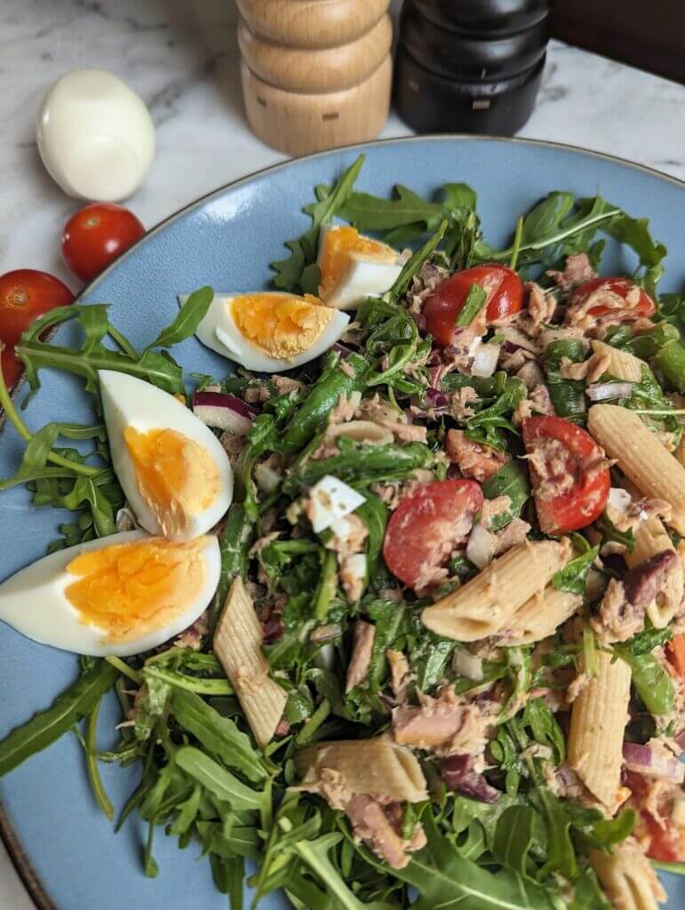 Pasta Niçoise on a plate next to salt, pepper, tomatoes and an egg