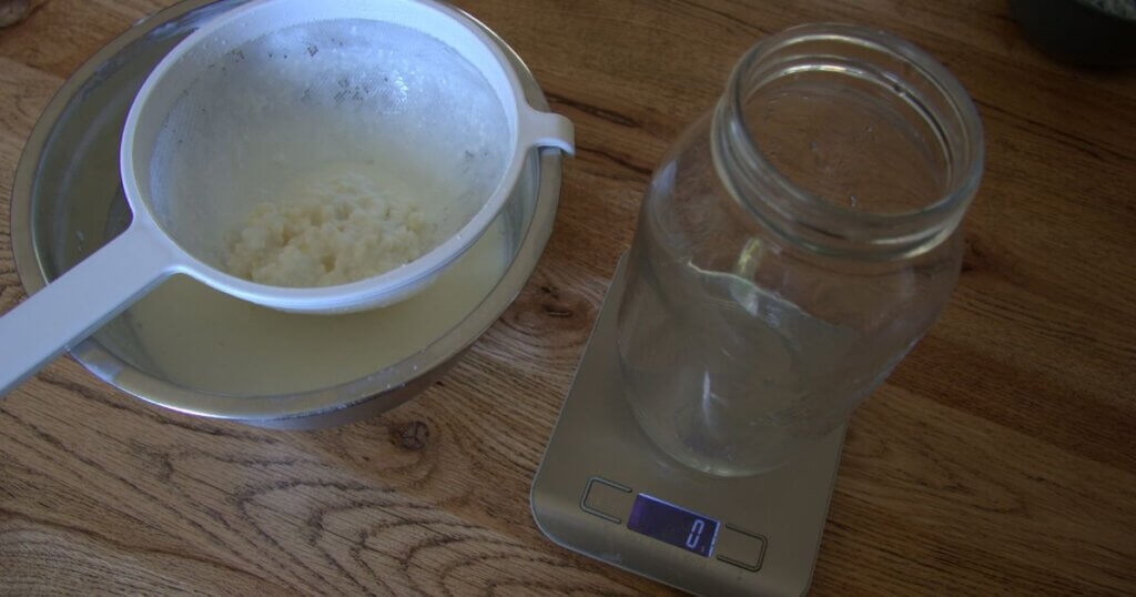 Straining milk kefir using a white plastic sieve over a metal bowl, with kefir grains visible in the strainer and an empty glass jar on a digital kitchen scale reading 0 grams.