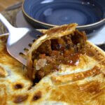 Beef pie with a spatula holding a piece out of the pie and a plate with knife and fork next to it on a wooden table