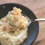 A spoon with rice pudding topped with cinnamon with a bowl in the background on a wooden table