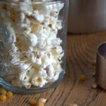 A glass jar filled with freshly popped popcorn sitting on a wooden surface, with scattered unpopped kernels and a metal measuring cup nearby.