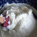 Close-up of creamy homemade cream cheese in a blue speckled bowl, with a red silicone spatula partially submerged.