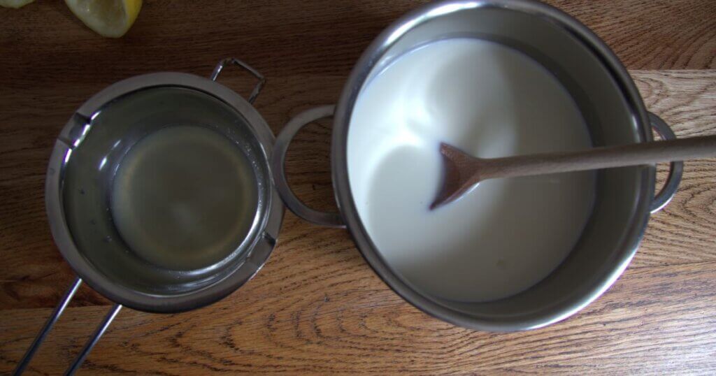 Overhead view of a pot of milk with a wooden spoon and a metal strainer containing freshly squeezed lemon juice, set on a wooden surface.