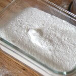 Close-up of freshly made powdered sugar in a glass container, with a blender lid and mixing jar dusted with sugar in the background on a wooden surface.