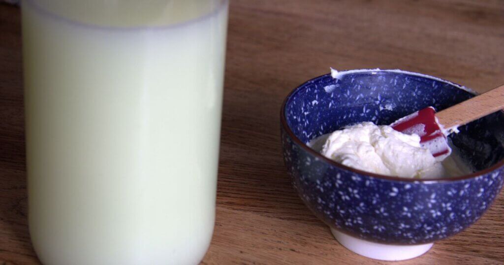 Jar of leftover whey next to a bowl of freshly made cream cheese with a red spatula on a wooden surface.