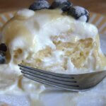 Close-up of a yogurt breakfast muffin topped with whipped cream, blueberries, and a drizzle of maple syrup, served on a striped plate with a fork.