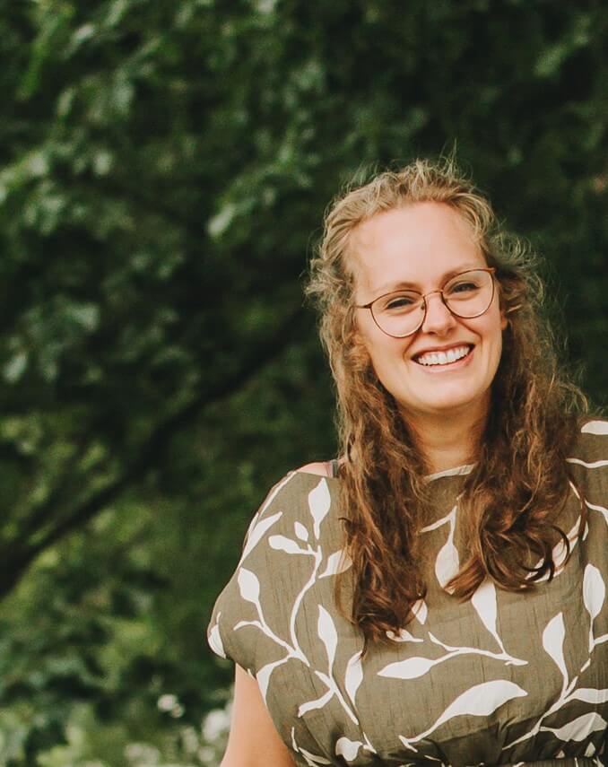 Smiling woman with glasses and long wavy hair, standing outdoors in front of a green leafy background, wearing a patterned top.